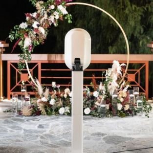 The white Ring Light Photobooth on its stand beside a floral arch at an outdoor reception