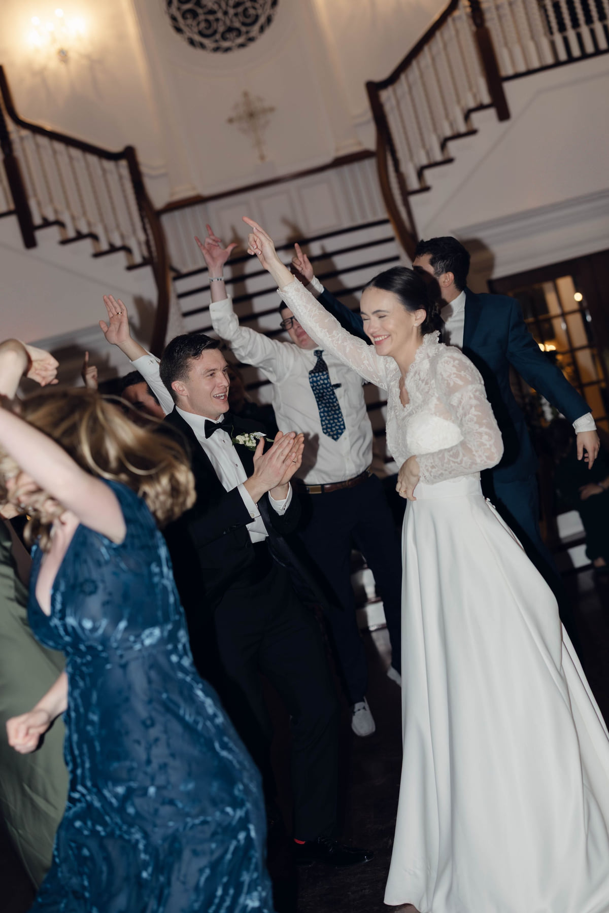 Bride in a lace gown laughing with one hand raised on a crowded dance floor