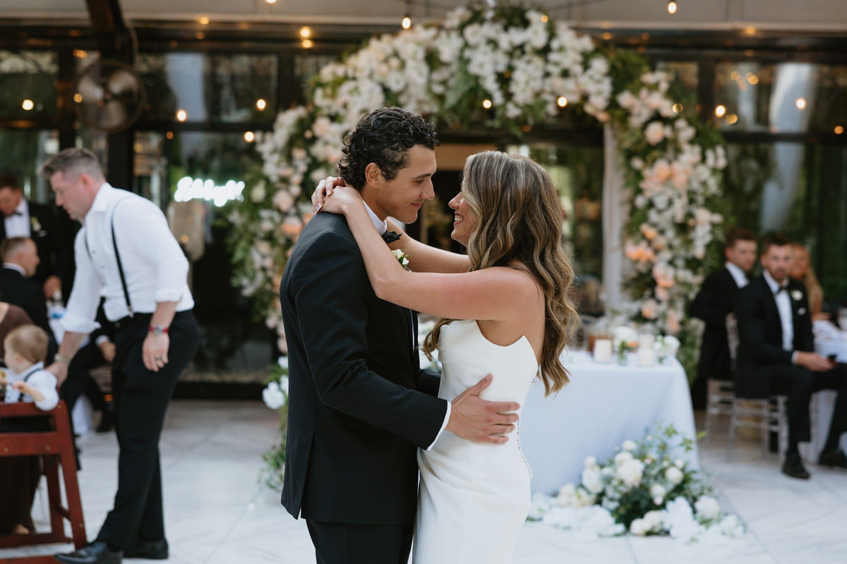 A couple in a first dance in front of a floral arch as seated guests watch