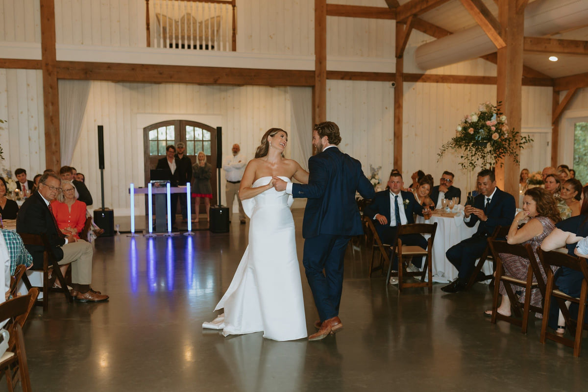 A couple dancing alone on an open floor in a bright barn as seated guests watch
