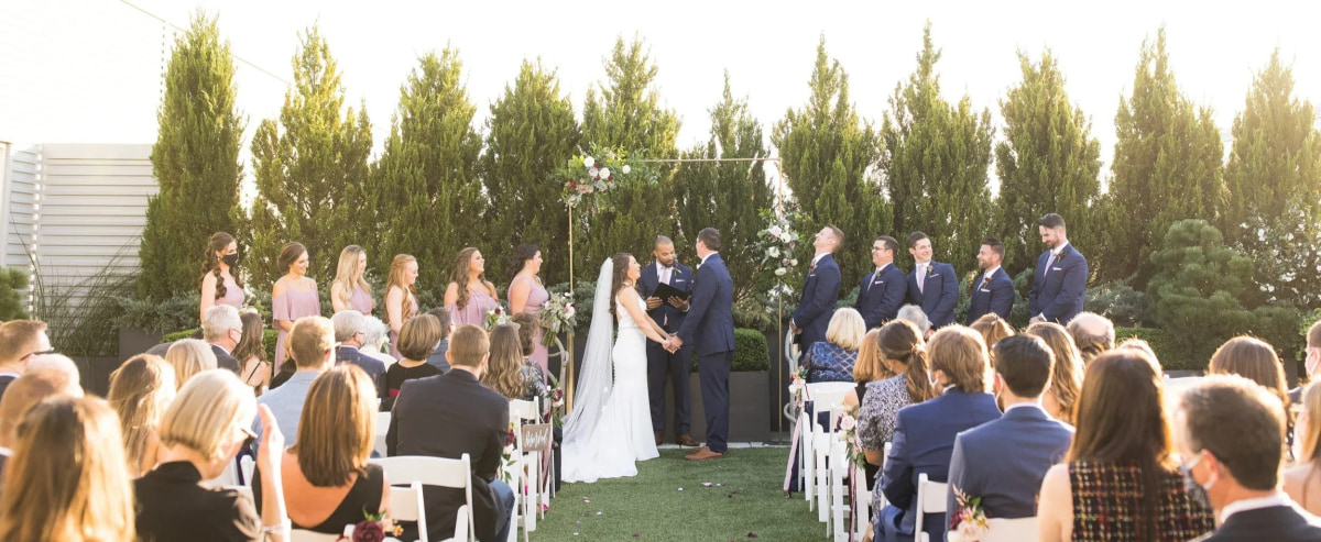 An outdoor wedding ceremony seen from behind the seated guests, under tall trees