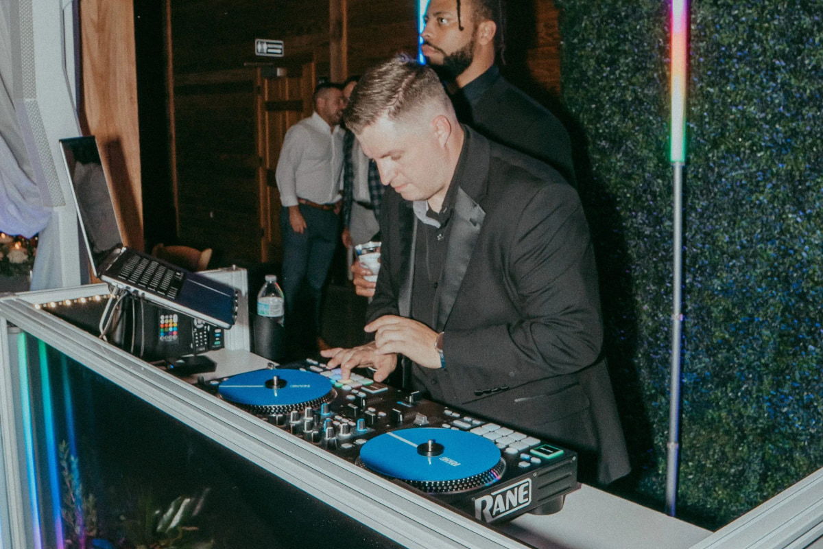 A DJ working a controller behind a white booth facade beside a lit hedge wall