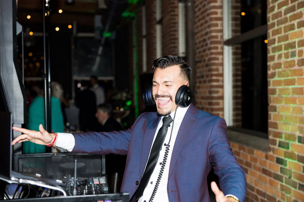 A DJ in a blue suit and headphones smiling at the decks in a brick room strung with lights
