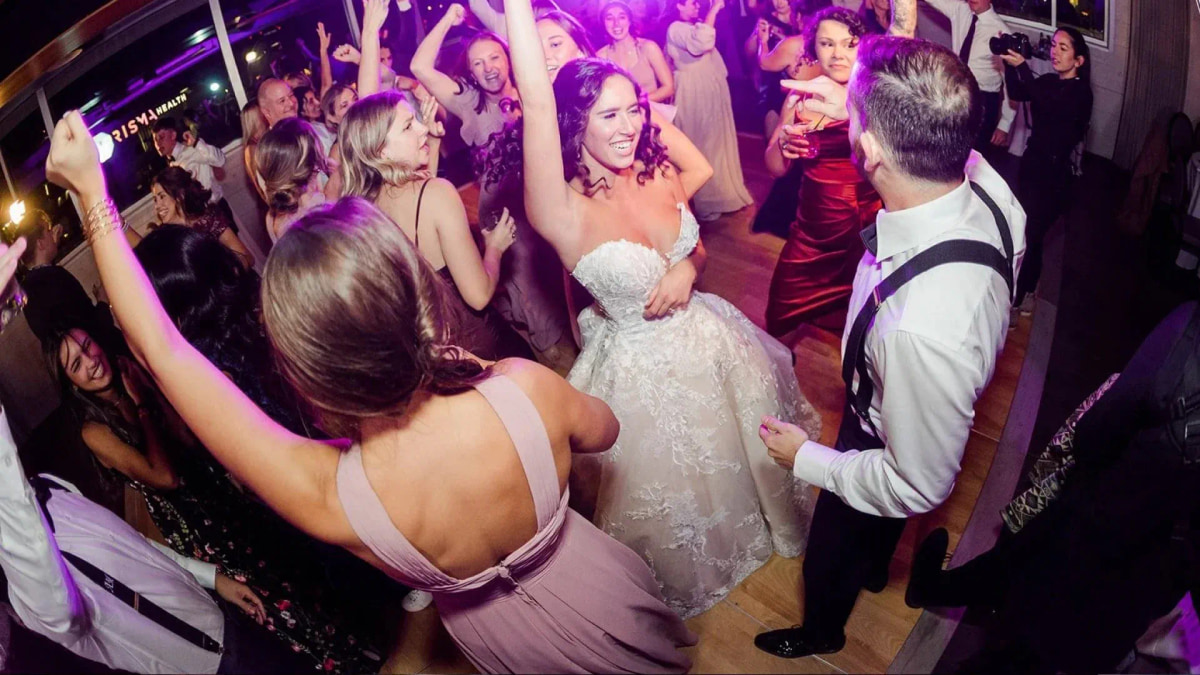 A packed wedding dance floor under purple light, the bride dancing with guests, arms in the air