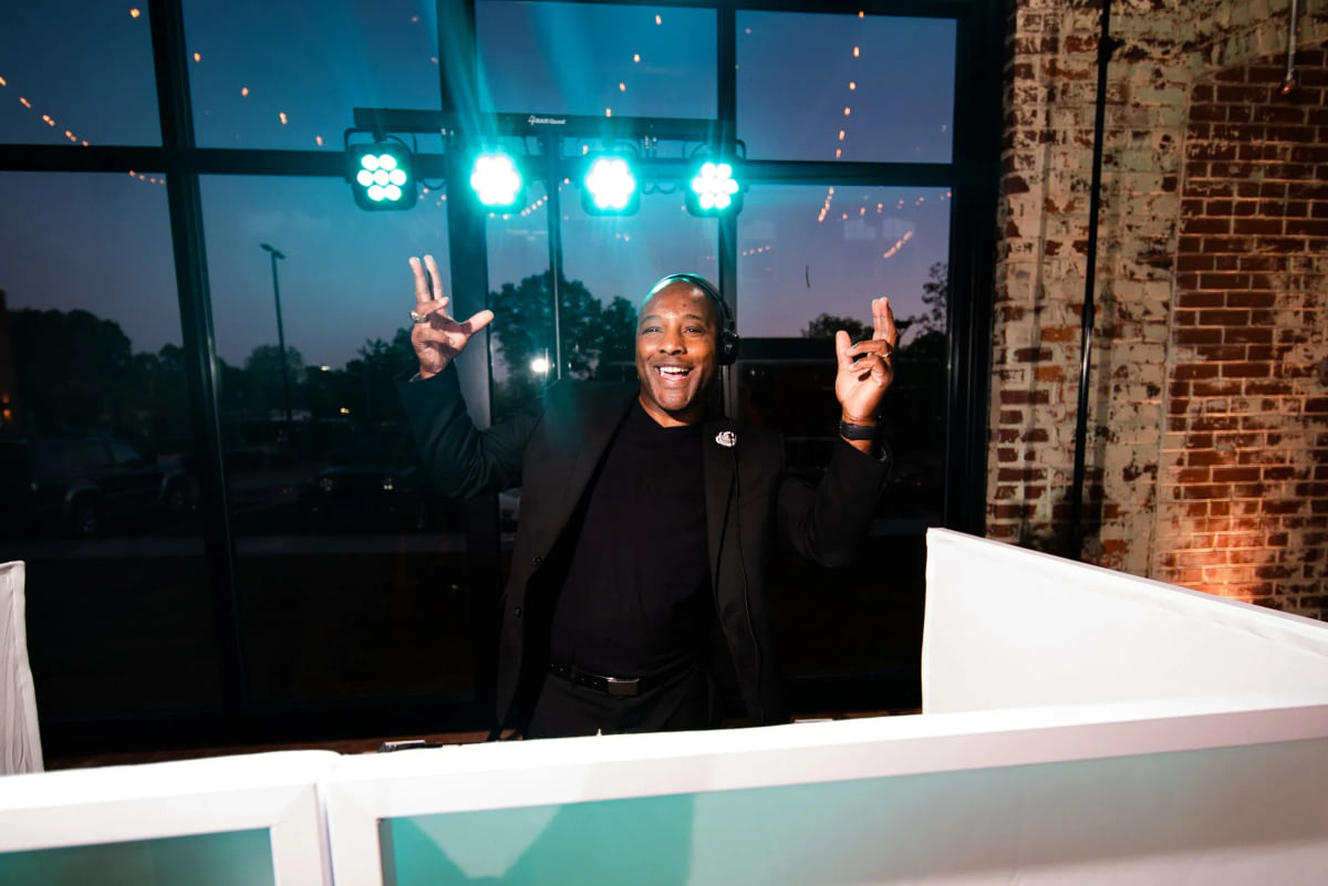 A DJ behind a white booth facade with both hands raised in front of a wall of windows at night