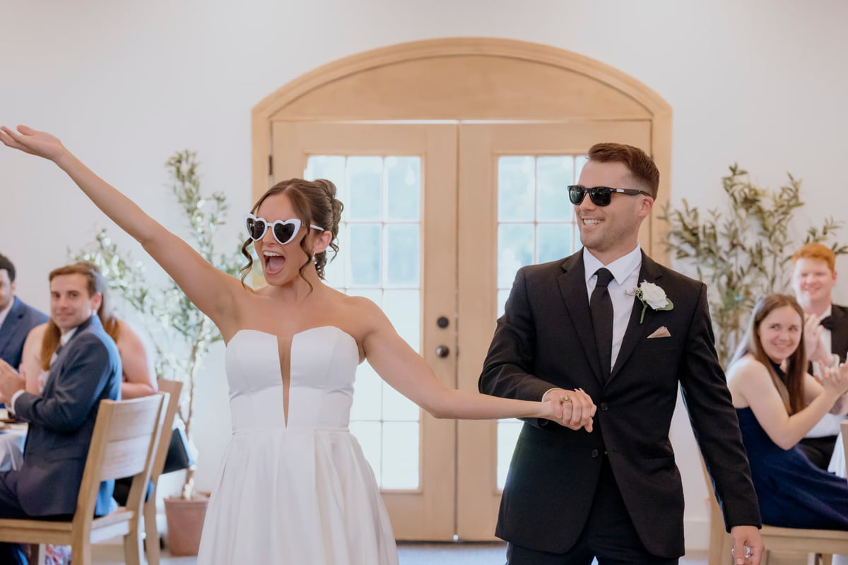 A bride in heart-shaped sunglasses throwing an arm in the air as she and the groom enter their reception to applause