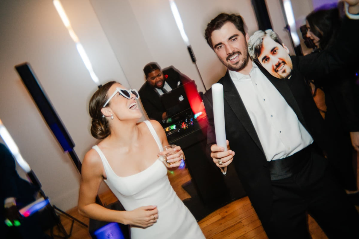 A bride in heart-shaped sunglasses laughing beside the groom on the dance floor while their SoundChoice DJ works the decks behind them
